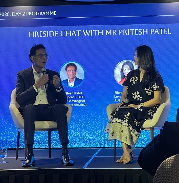 Pritesh Patel and Richa Goyal Sikri seated on stage during a fireside chat, with a screen behind them reading "Fireside Chat with Mr Pritesh Patel.
